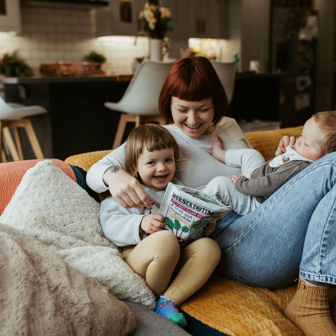 A white woman, with dark brown short bob sits on a rug in a living room. On her lap is a small toddler with a dummy in her mouth. Lying on the floor, looking up at the mother is a small baby.