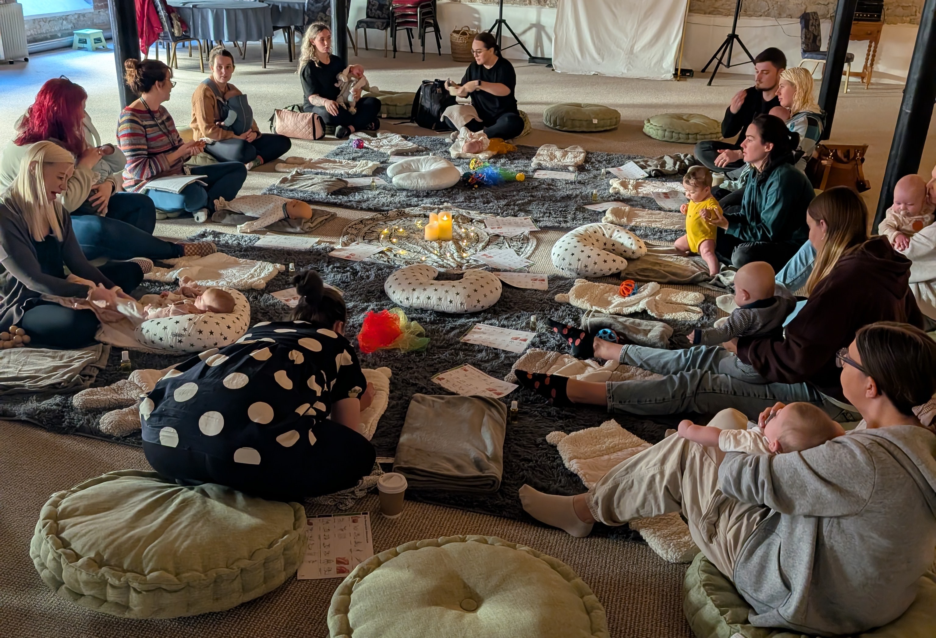 A group of parents with their babies sit on the floor, in a large circle. On the floor are toys and cushions for babies.