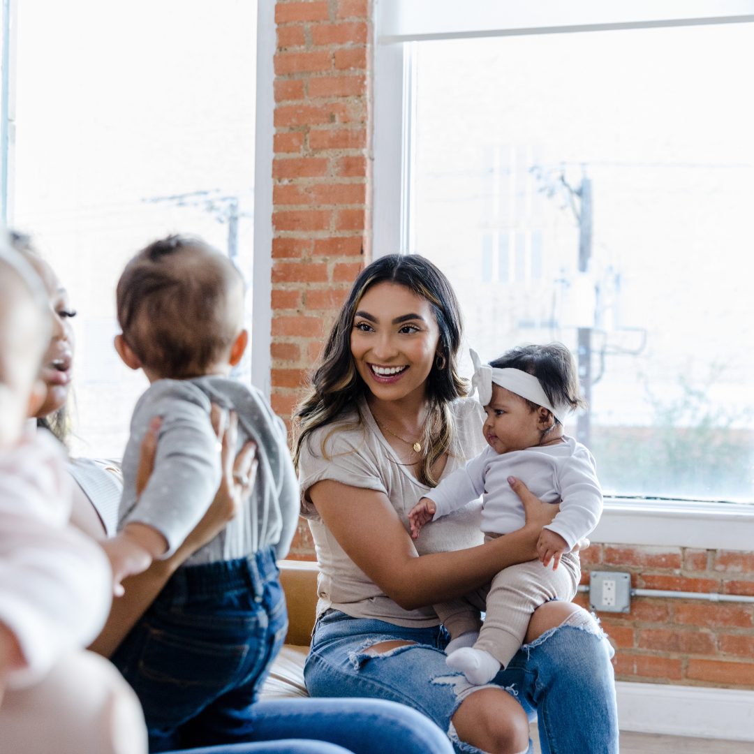 A white woman, with dark brown short bob sits on a rug in a living room. On her lap is a small toddler with a dummy in her mouth. Lying on the floor, looking up at the mother is a small baby.